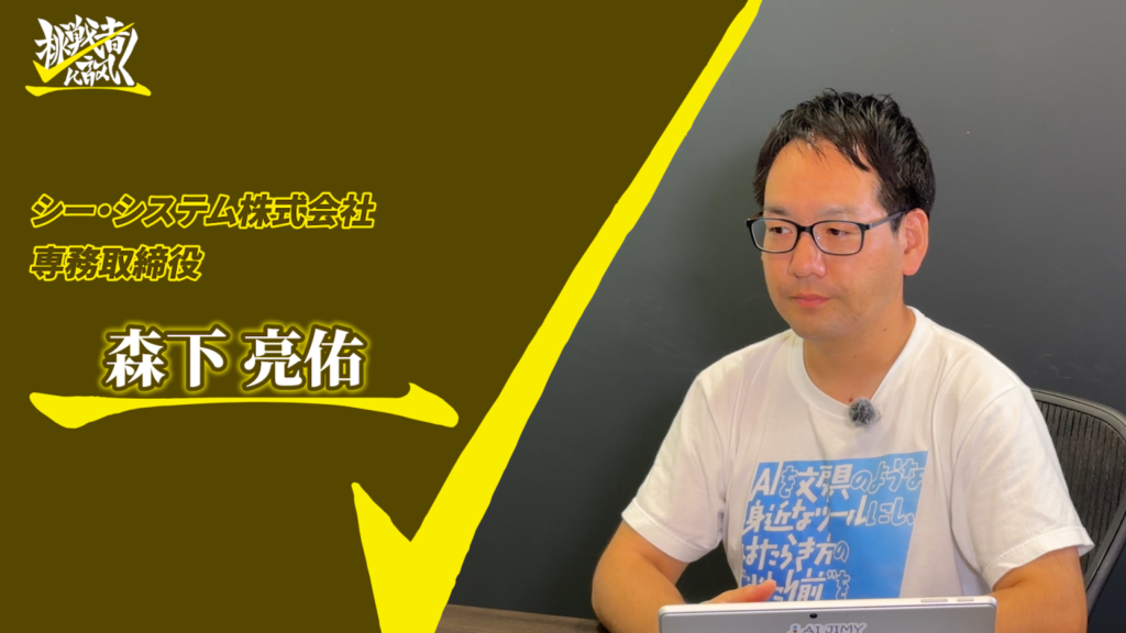 Portrait of 森下亮佑 wearing glasses and a white T‑shirt, seated at a desk; brown left panel with yellow Japanese text and a logo is visible.